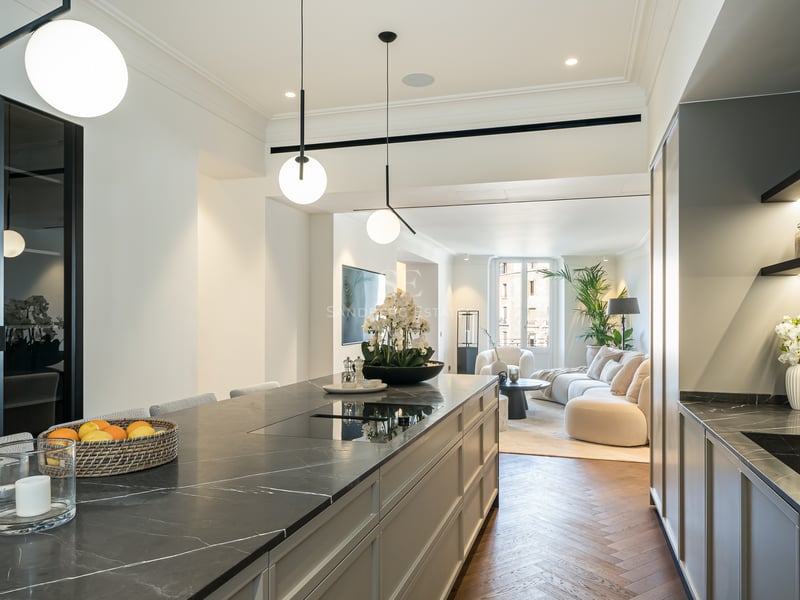 Modern kitchen featuring a dark marble island and herringbone wood floors, opening into a bright living room area.