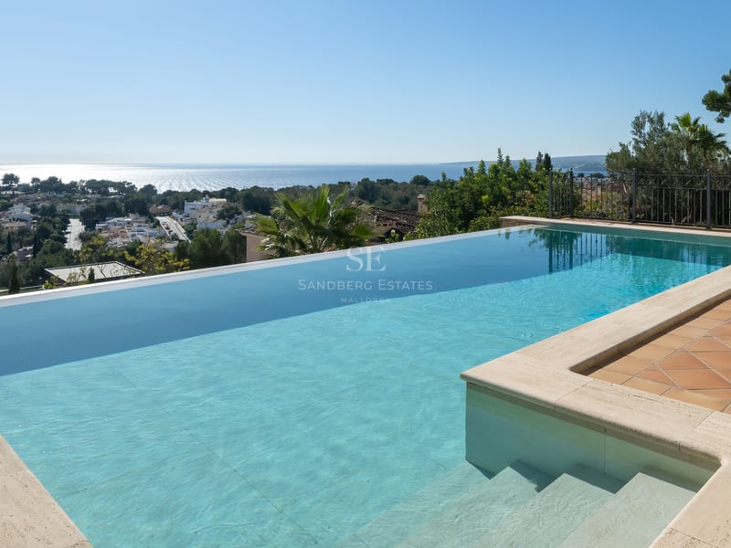 Piscine à débordement en L avec margelles en travertin et terrasse en terre cuite dominant le littoral et la mer.