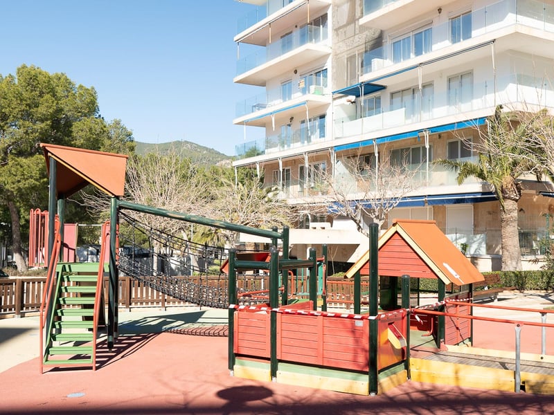 Aire de jeux pour enfants avec structures en bois et sol sécurisé devant un immeuble moderne.