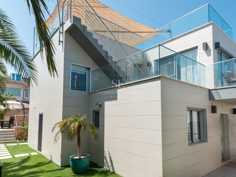 Façade d'une villa moderne avec garde-corps en verre, escalier extérieur, voile d'ombrage et jardin paysager.