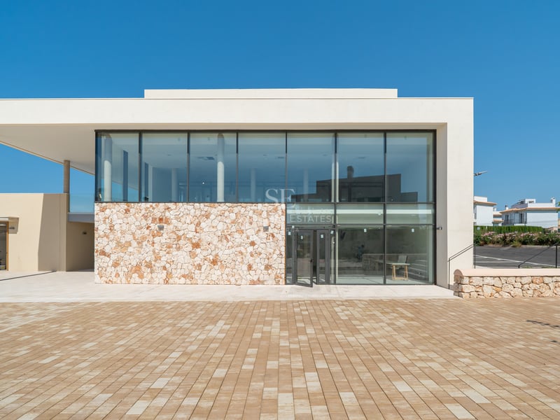 Fachada de edificio moderno con grandes ventanales de cristal, muro de piedra natural y un amplio patio pavimentado.
