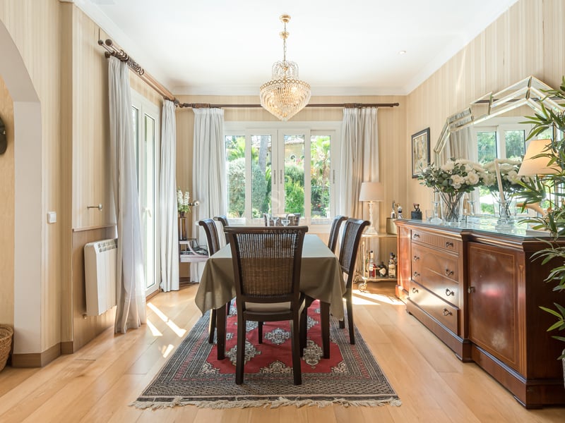 Salle à manger ensoleillée avec table en bois, lustre en cristal et grand buffet donnant sur l'extérieur.