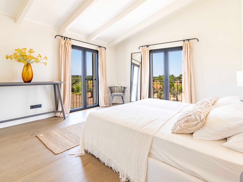 Bright master bedroom featuring white vaulted ceilings with exposed beams, two sets of dark-framed French doors, and oak flooring.