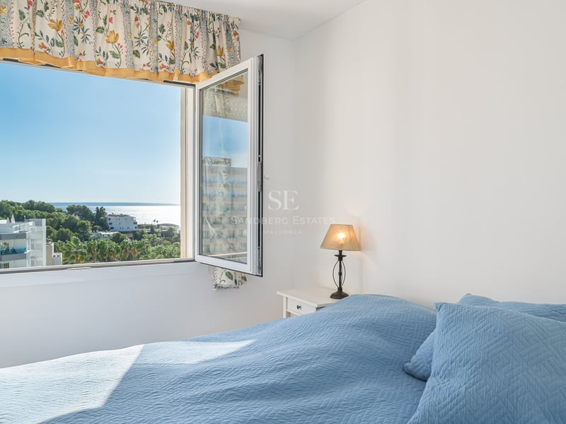 Chambre avec lit bleu et fenêtre ouverte offrant une vue sur un paysage côtier avec mer et arbres.