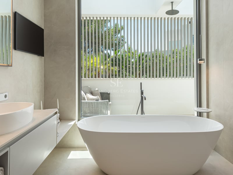 Salle de bains minimaliste avec baignoire îlot blanche, vasque à poser et grande fenêtre sur terrasse privée ensoleillée.