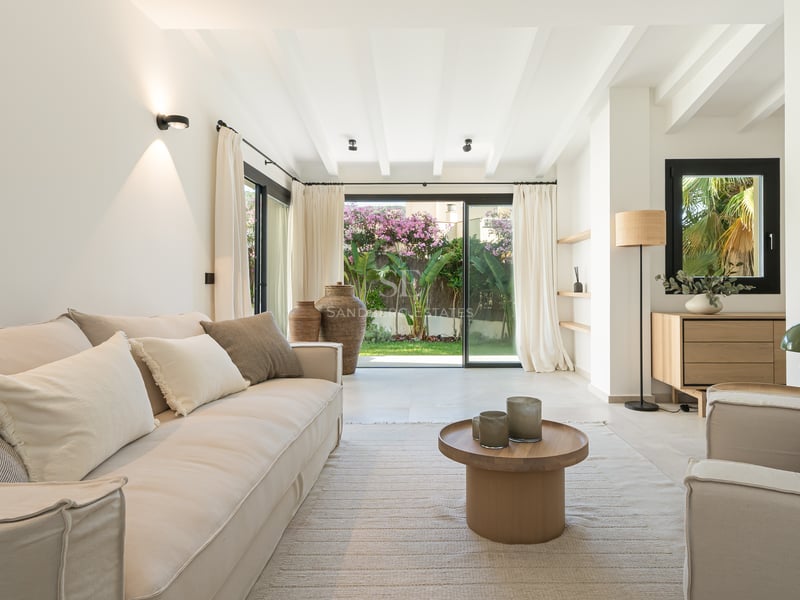 Salon lumineux avec canapés beiges et grandes baies vitrées ouvrant sur un jardin privé.