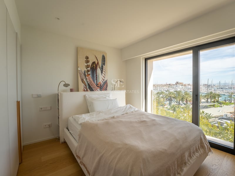 Chambre moderne avec lit blanc et grande fenêtre offrant une vue sur une marina ensoleillée avec des palmiers.
