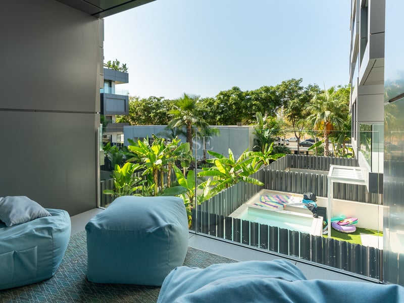 Balcon moderne avec poufs bleu clair et garde-corps en verre surplombant un jardin tropical et une piscine.