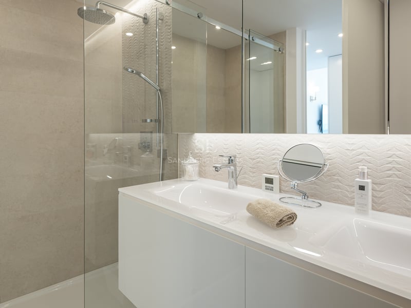 Salle de bain contemporaine avec double vasque blanche, douche vitrée et carrelage beige texturé.