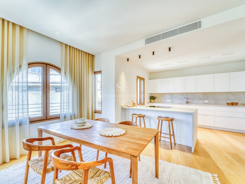 Bright open-plan kitchen with white cabinetry and a wooden dining table under an arched window with light curtains.