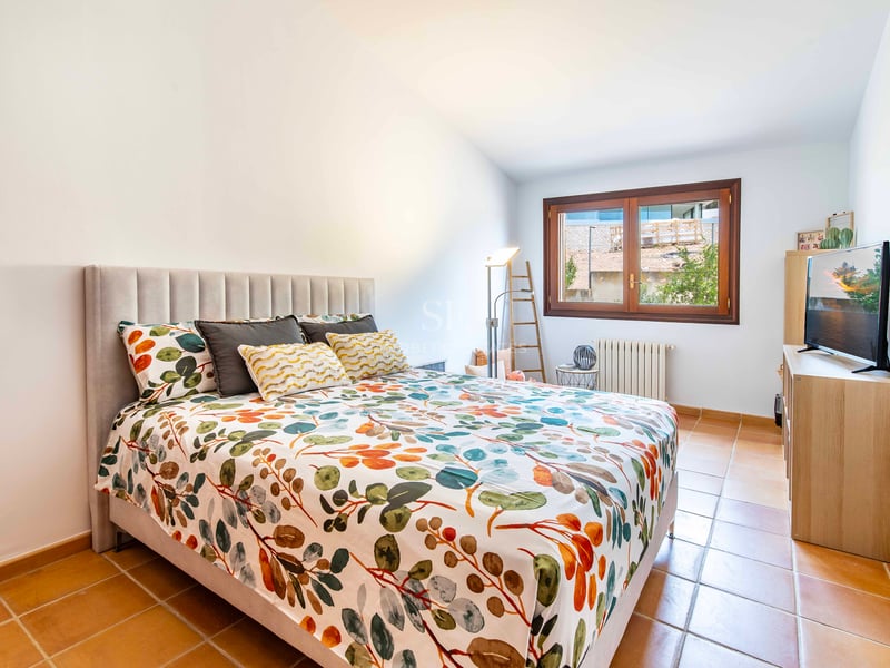 Chambre avec lit à motifs floraux, tête de lit grise, sol en terre cuite et fenêtre en bois.