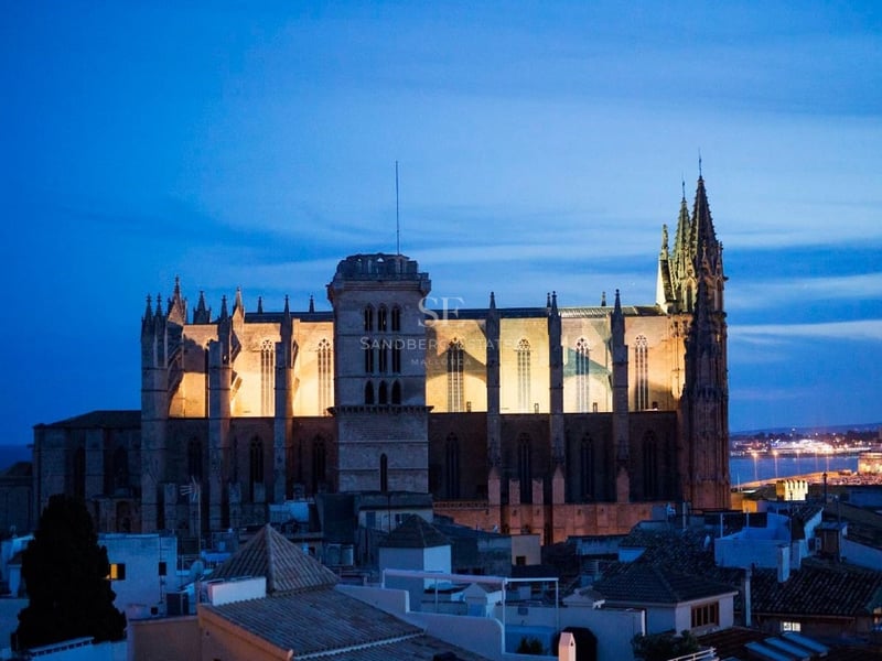 Majestuosa catedral gótica iluminada con luz cálida bajo un cielo azul profundo de noche sobre tejados urbanos.