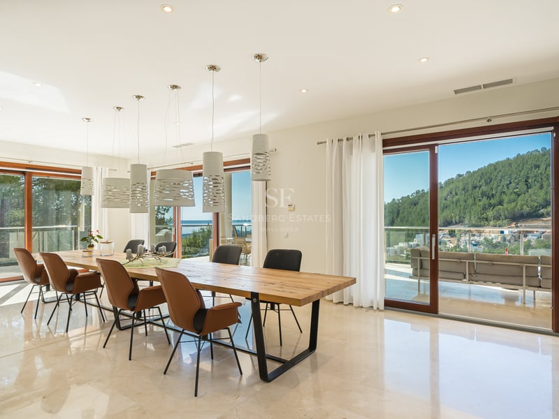 Salle à manger moderne avec table en bois, sol en marbre et baies vitrées ouvrant sur une vue montagne et mer.