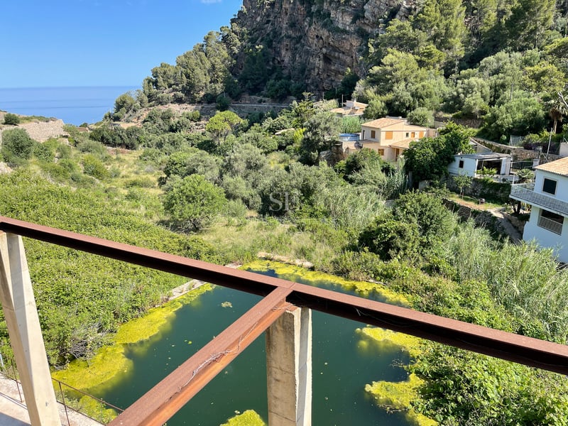 Vue depuis une terrasse en acier surplombant une vallée verdoyante avec des maisons méditerranéennes et des falaises.