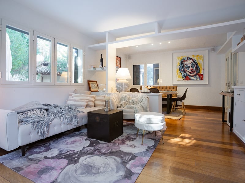 Salon à aire ouverte avec canapés blancs, tapis floral, parquet en bois et coin repas en arrière-plan.
