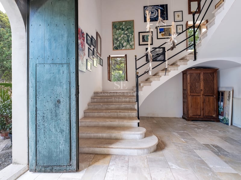 Hall d'entrée avec une grande porte bleue vieillie, escalier en pierre et galerie de tableaux sur murs blancs.