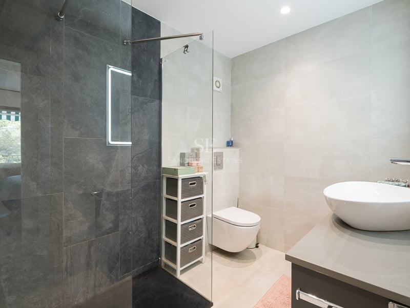 Salle de bain moderne avec douche en verre, carrelage gris foncé, vasque blanche et toilettes suspendues.