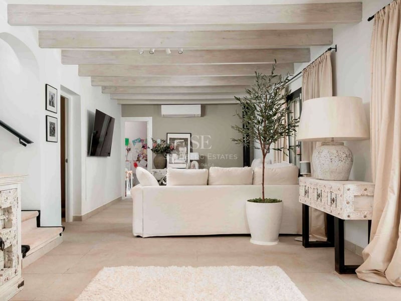 Salon lumineux avec canapé blanc, poutres en bois clair, sol en pierre et grande baie vitrée.