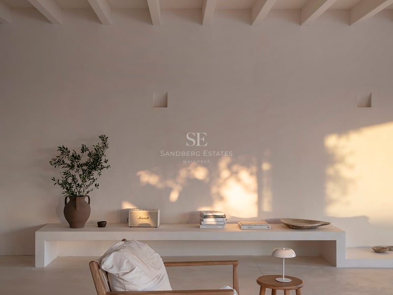 A minimalist room with white beamed ceilings, a wooden armchair, and warm golden hour light hitting a plastered wall.