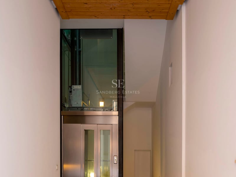 Couloir moderne avec sol en mosaïque de pierre, plafond en bois et ascenseur en verre au fond.