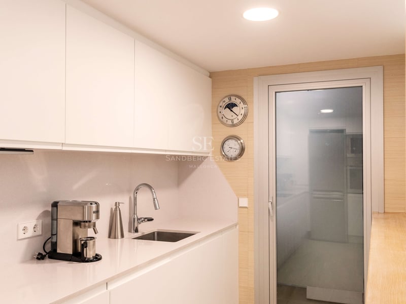 Cuisine en couloir blanche avec placards sans poignées, évier en inox et sol en bois clair.
