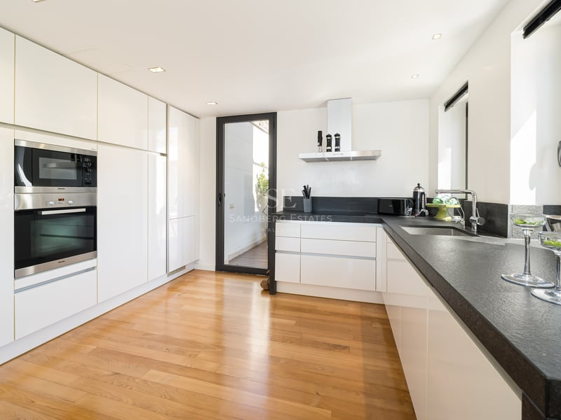 Cuisine moderne blanche avec plans de travail en pierre sombre, parquet en chêne, appareils intégrés et accès terrasse.