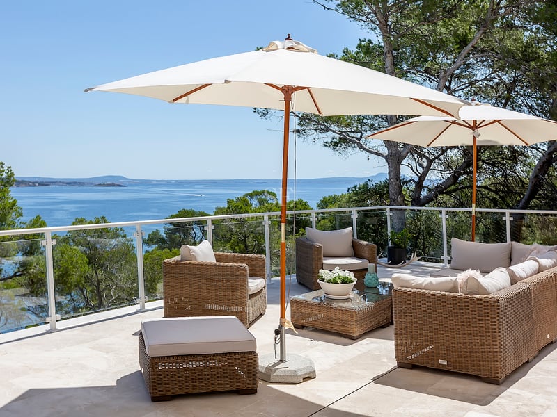 Terrasse ensoleillée avec salon en rotin, parasols blancs et garde-corps en verre surplombant la mer bleue.