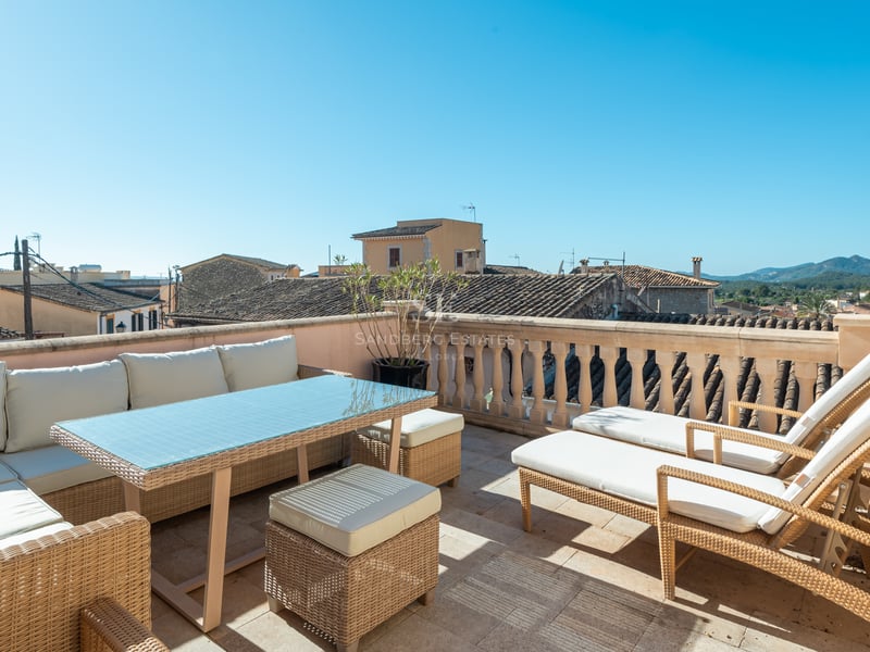Terrasse ensoleillée avec mobilier en rotin, coussins blancs et vue sur les toits historiques et les collines.