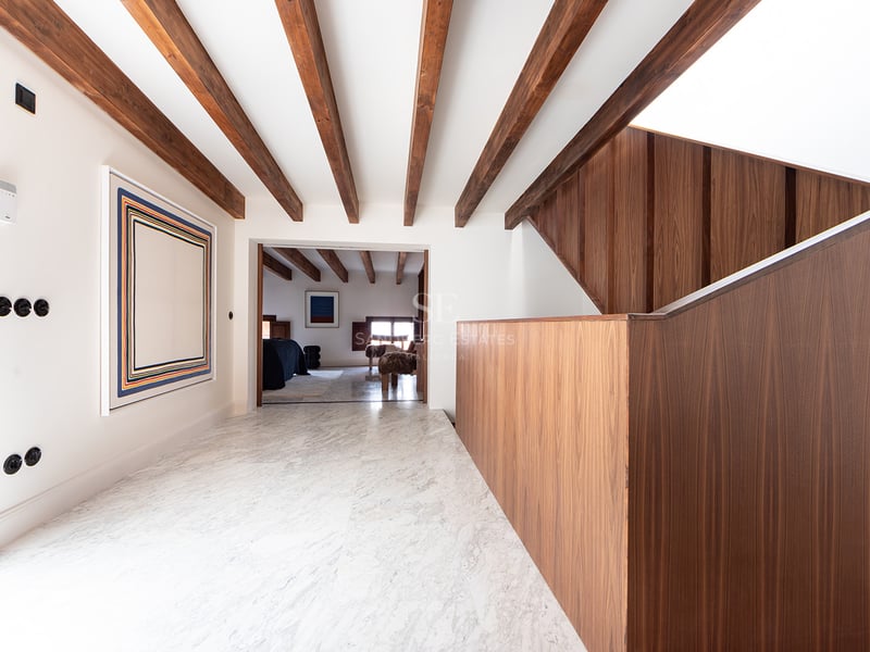 Pasillo luminoso con suelos de mármol blanco, vigas de madera oscura en el techo y una escalera moderna de madera.