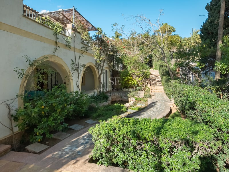 Sentier en pierre à travers un jardin méditerranéen luxuriant à côté d'une villa blanche aux arcades.