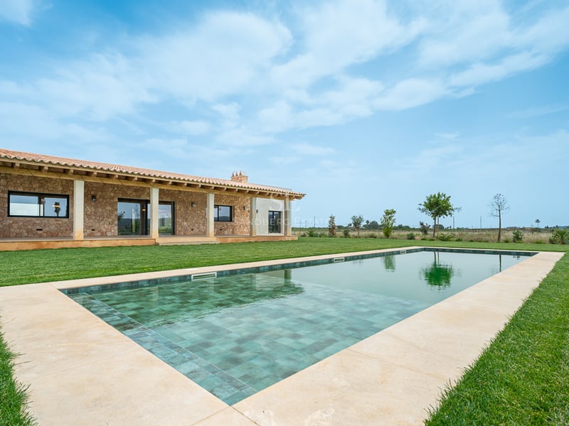Grande piscine rectangulaire aux carreaux turquoises face à une villa traditionnelle en pierre et pelouse verte.