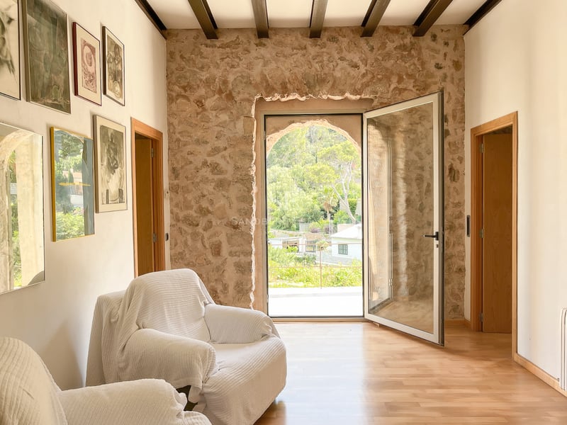 Intérieur d'un salon avec mur en pierre naturelle, poutres en bois et porte vitrée donnant sur un jardin verdoyant.