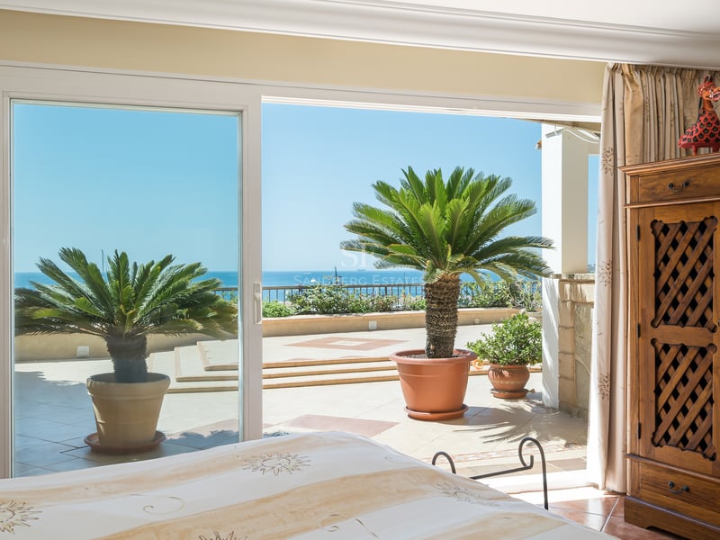 Chambre principale lumineuse avec armoire en bois et baies vitrées ouvrant sur une terrasse avec palmiers et vue mer.