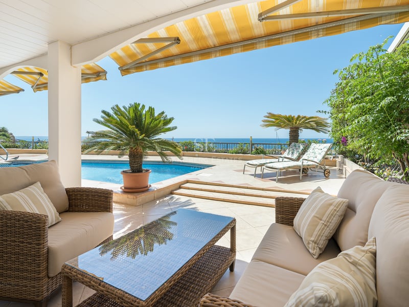 Terrasse couverte avec canapés en osier face à une piscine bleue et la mer sous un ciel dégagé.