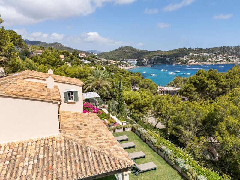 Vue en plongée d'une villa au toit de tuiles surplombant un jardin, une baie turquoise avec des bateaux et des collines.