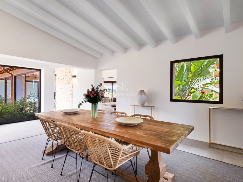 Salle à manger lumineuse avec table en bois, chaises en osier et plafond à poutres blanches.