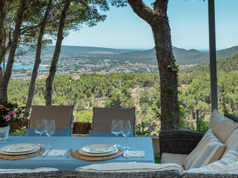 Table à manger extérieure et canapé en osier sur une terrasse ombragée surplombant une ville côtière et la mer.