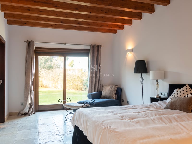 Chambre spacieuse avec poutres apparentes en bois, sol en pierre et baie vitrée ouvrant sur un jardin.