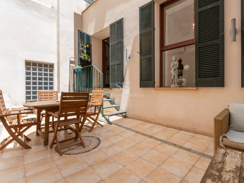 Terrasse privée carrelée avec table à manger en bois, volets verts et salon extérieur en plein jour.