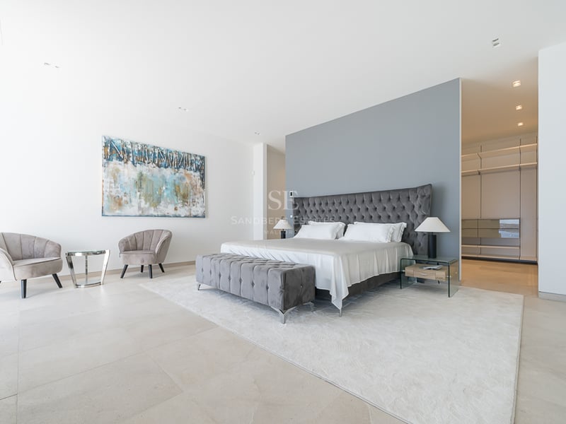 Chambre principale avec lit capitonné gris, coin salon en velours et aperçu d'un dressing.