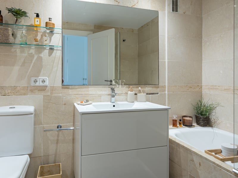 Salle de bain lumineuse avec carrelage en marbre beige, meuble vasque blanc, grand miroir et baignoire intégrée.