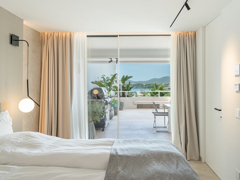 Chambre moderne avec lit blanc et portes vitrées s'ouvrant sur une terrasse avec barbecue et vue sur la mer.