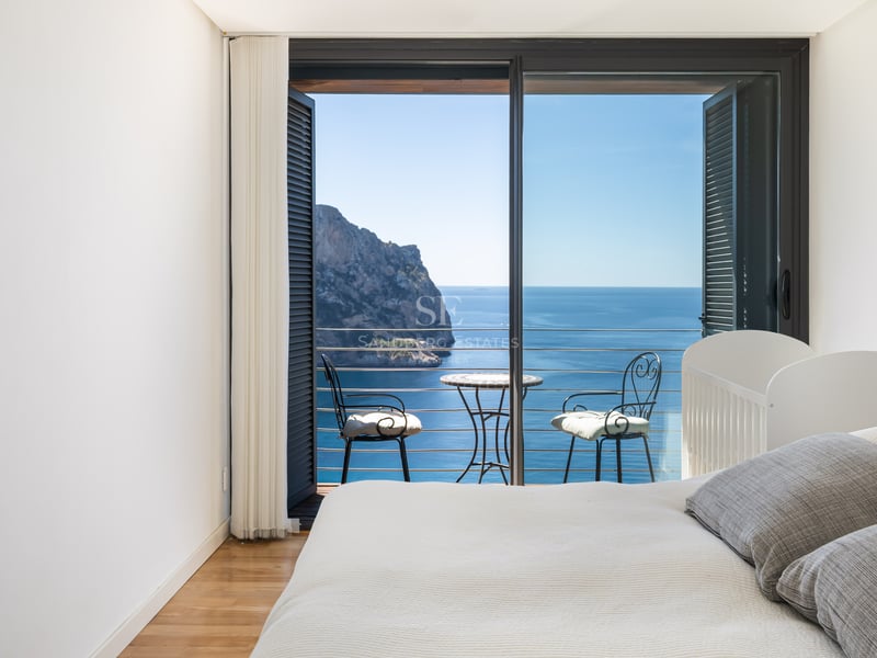 Chambre minimaliste blanche avec balcon privé, mobilier en fer forgé et vue sur une falaise et la mer Méditerranée.