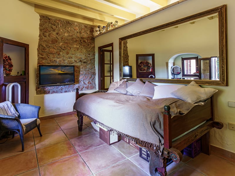 Une chambre chaleureuse avec un sol en terre cuite, un mur en pierre avec TV et un grand miroir au-dessus du lit.