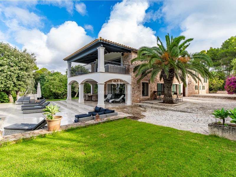 Vue extérieure d'une villa en pierre avec arches blanches, un grand palmier, des transats et un jardin paysager.