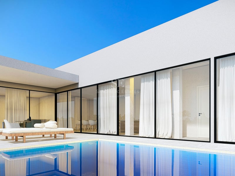 Villa blanche moderne avec de grandes baies vitrées surplombant une piscine bleue et une terrasse en pierre avec transats.
