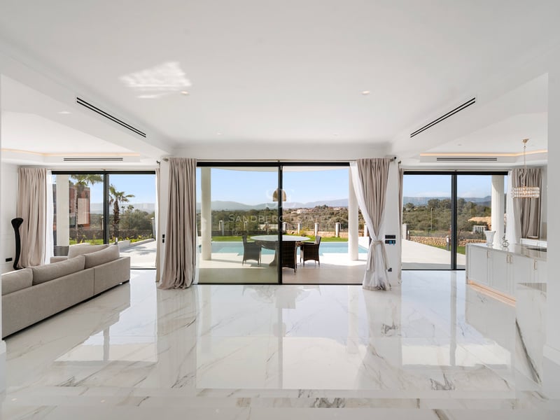 Salon lumineux avec sol en marbre blanc et baies vitrées donnant sur une piscine et les collines.
