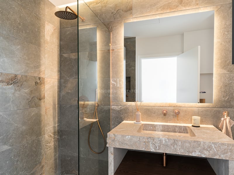 Salle de bains de luxe avec douche à l'italienne, vasque en pierre et grand miroir rétroéclairé avec lumière chaude.