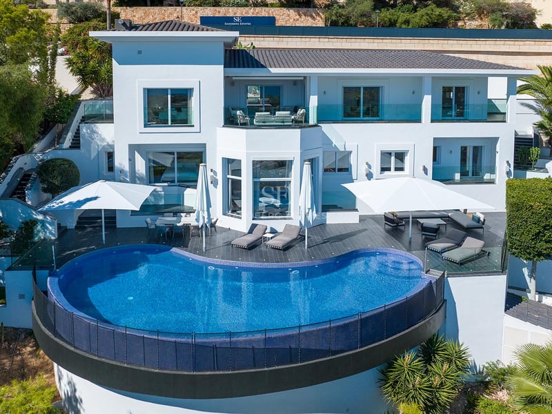 Vue aérienne d'une villa blanche moderne avec une grande piscine à débordement bleue et des terrasses meublées.