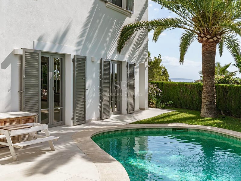 Piscine turquoise à côté d'une villa blanche avec un palmier, des volets gris et une vue lointaine sur l'océan.